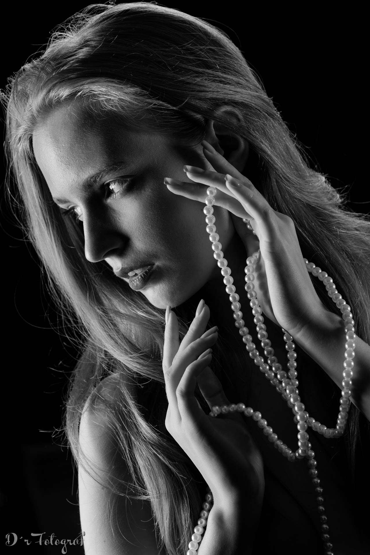 Black-and-white portrait of a woman with long, flowing hair, gazing to the side. She is holding a string of pearls delicately between her fingers. The lighting creates dramatic shadows, highlighting the contours of her face and hands. The background is dark, emphasizing the subject and the texture of her skin and hair. The image has an artistic and elegant feel.