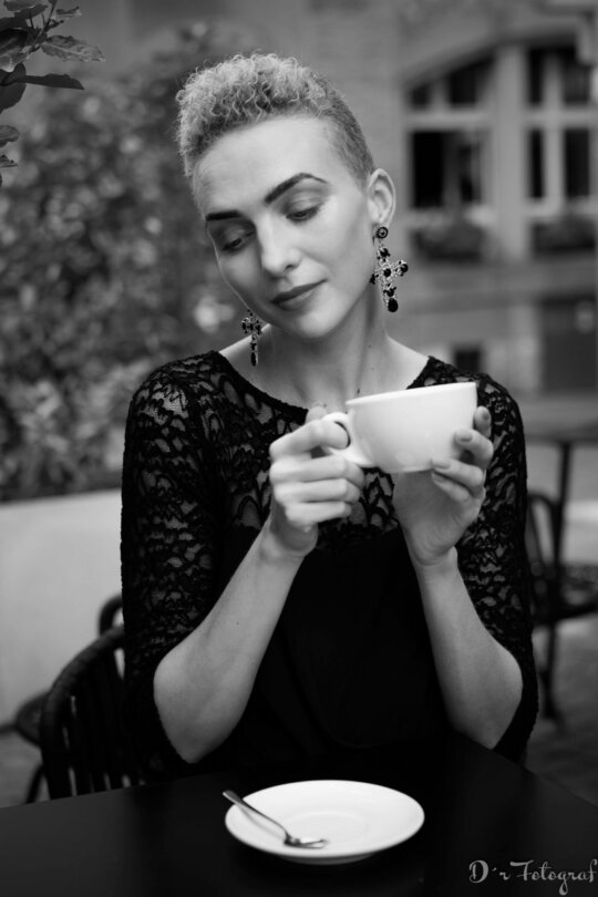 Frau mit kurzen Haaren in schwarzer Spitzenbluse hält grosse weisse Kaffeetasse in der Hand, sitzt an einem Kaffeetisch im Freien und schaut verträumt  nach unten